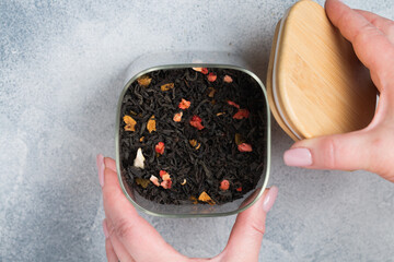 Black leaf tea with fruit additives (strawberry, raspberry, apple, pear, feijoa) in a glass jar in women's hands on a gray background. A delicious and healthy drink. Selective focus, copy space