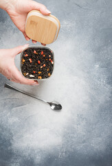 Black leaf tea with fruit additives (strawberry, raspberry, apple, pear, feijoa) in a glass jar in women's hands on a gray background. A delicious and healthy drink. Selective focus, copy space