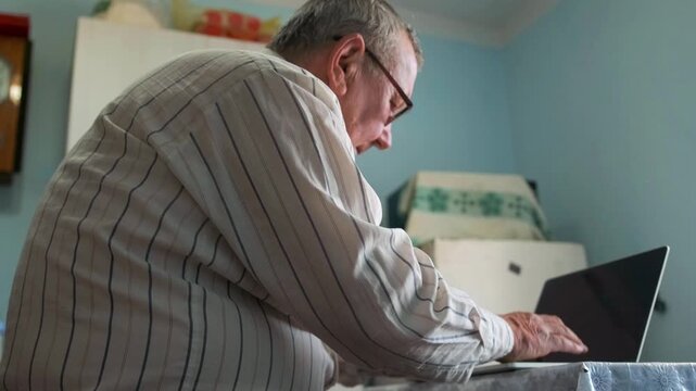 Elderly Man Uses Computer Diligently - Mature Caucasian Gentleman Intently Focused On His Digital Task - An Aged Caucasian Man Attentively Engaged In Typing On His Laptop In Modest Kitchen