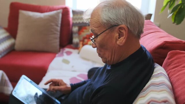 Elderly Man Flipping Through Pictures Displayed On Tablet Screen In Cozy Living Room - Older Man Leisurely Exploring His Photograph Collection Displayed On Digital Tablet In Comfortable Sitting