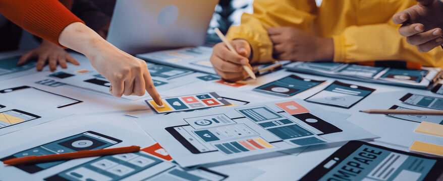 A diverse team engages in a collaborative design workshop, reviewing mobile app wireframes and discussing creative ideas over a table filled with prototypes and tools. SACTR - Powered by Adobe