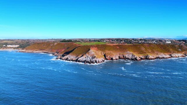 Sloping rocky coast surrounded by the blue waterscape. Residential area scattered on the picturesque shore of Wales, Great Britain.