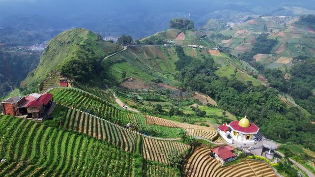 Aerial drone footage of an agricultural valley with green hills, a gold roof mosque, a countryside road, in the mountain touristic spot of Panyaweuyan, Majalengka regency, Java island, Indonesia