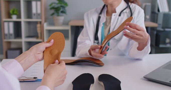 Young friendly female orthopedist doctor sitting at the desk in medical clinic showing orthopedic insoles to woman patient and consulting about feet health. Medicine and orthopedics concept. 4k video.