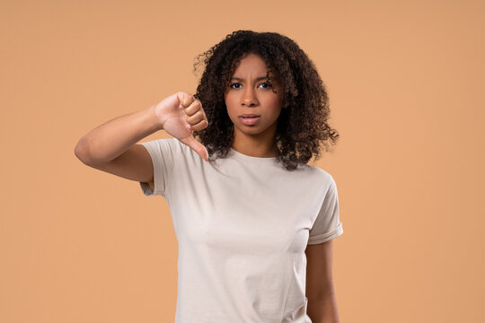 African Woman With Dislike Sign. Pretty Lady Showing Thumbs-Down on beige studio