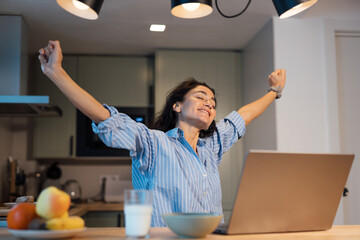 Happy young woman in striped pajamas stretching arms with closed eyes at kitchen table while working on laptop, relaxed morning routine, remote work lifestyle, comfort, freedom, positive emotion