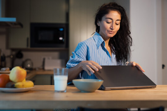 Happy young woman in striped pajamas stretching arms with closed eyes at kitchen table while working on laptop, relaxed morning routine, remote work lifestyle, comfort, freedom, positive emotion - Powered by Adobe