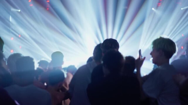 Audience stands shoulder to shoulder facing concert stage as powerful strobe lights radiate outward creating immersive live music atmosphere