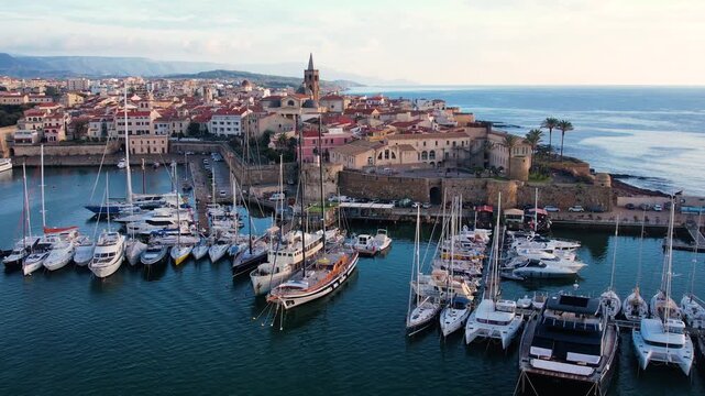 Enjoy stunning aerial views of the marina in Alghero, Sardinia, featuring boats and yachts against a backdrop of historic architecture and the beautiful coastline.