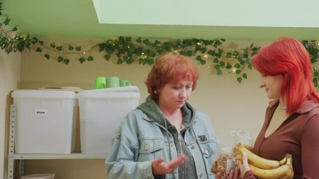 caucasian women in pantry exchanging groceries, older coordinator stocks shelves while younger donor offers bananas and granola warm lighting and ivy decor create friendly volunteer atmosphere, denim