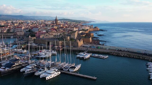 Enjoy stunning aerial views of the marina in Alghero, Sardinia, featuring boats and yachts against a backdrop of historic architecture and the beautiful coastline.