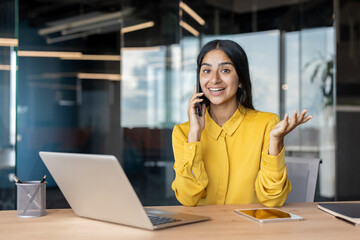 Smiling young businesswoman sitting at desk in office and talking on phone