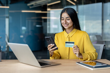 Smiling young Indian woman sitting in the office at a desk with a laptop, holding a credit card in...