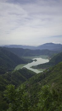 Vertical aerial view of a scenic green mountain valley with a turquoise lake under a cloudy sky