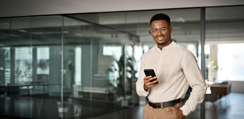 Smiling busy professional African American business man standing in office holding mobile...