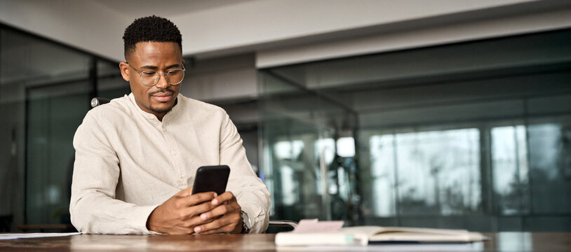 Busy African American entrepreneur using cell phone working in office. Professional young Black business man holding mobile cellphone looking at cellphone making financial bank payment at work.