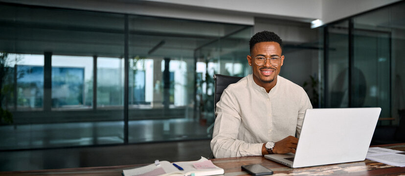 Portrait of smiling male company employee or entrepreneur in office. Happy young professional African American business man in glasses looking at camera sitting at work desk using computer.