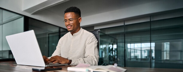 Busy smiling professional African business man company employee, young male worker businessman wearing glasses typing on computer using laptop working at office, elearning at workplace desk.