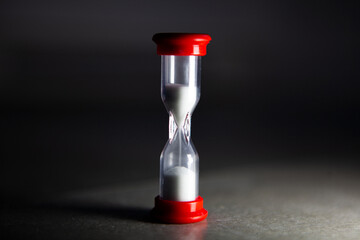 Minimal still life of a small hourglass with red caps, white sand flowing on a dark background, symbolizing time, deadline, countdown, urgency, productivity and focus.