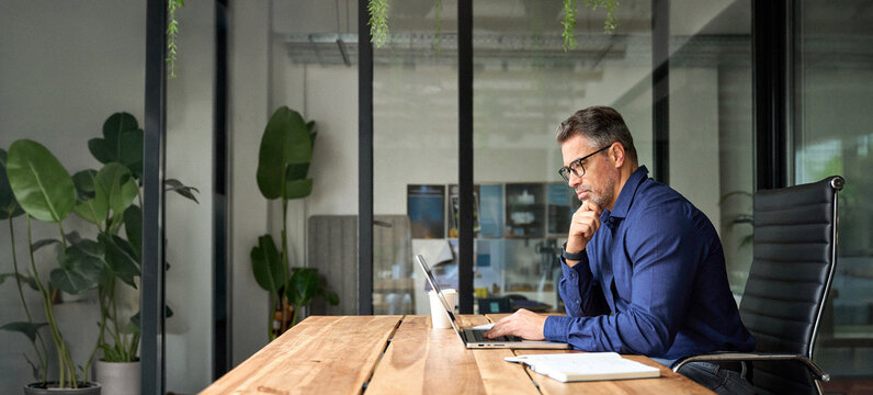 Busy business man of middle age using laptop sitting at office desk. Mature older professional businessman executive at work. 50 years old manager or investor working on computer technology.