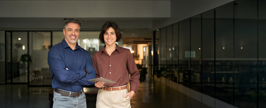2 smiling company managers, businessman and businesswoman leaders professional team standing at work looking at camera. Two happy executives business man and woman partners in office. Portrait.