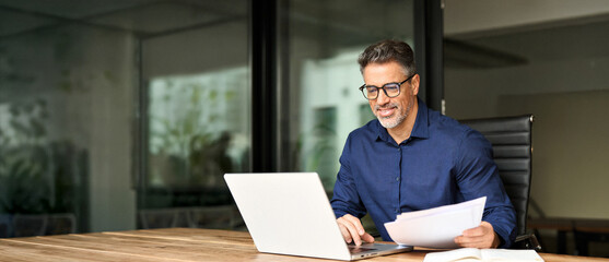 Happy busy older professional businessman entrepreneur using laptop holding bill corporate document...