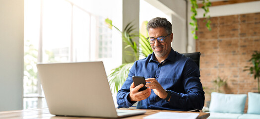 Happy middle aged businessman using mobile phone working in office. Busy smiling 50 years old man...