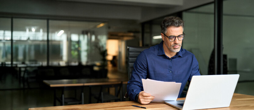 Middle aged professional business man using laptop computer reading document at work. Busy mature male account bank manager executive working on laptop checking financial report in office. Copy space.