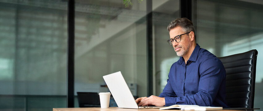 Busy business man of middle age using laptop sitting at office desk. Mature older professional businessman executive at work. 50 years old manager or investor working on computer technology.