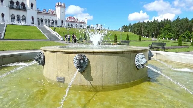 People walk around a fountain with lion heads, enjoying the sunshine and greenery in a large garden. The historic building The Puslovsky Palace in Kossovo stands in the background, showing rich archit