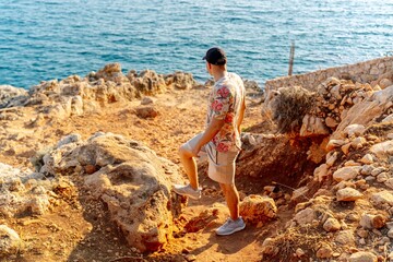 Fototapeta premium Young man standing on a rocky seaside cliff looking at the sea during summer travel