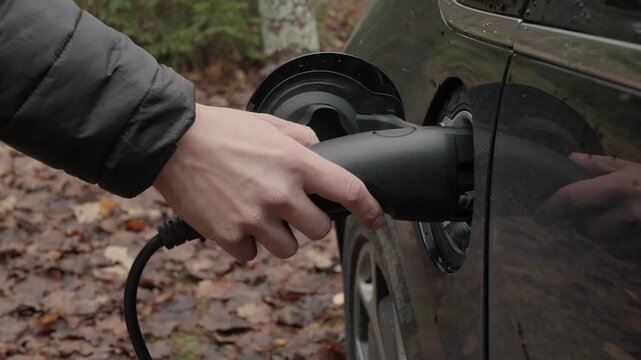 Driver plugs electric car charger outdoors in wet forest parking, hand inserts connector into vehicle port, autumn leaves on ground, overcast light, eco commute and sustainable