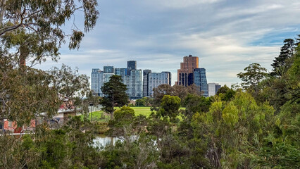 Modern city skyscrapers rise above a peaceful park and reflective pond, framed by lush green trees and natural foliage under a soft sky.