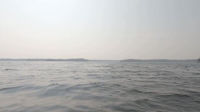 Point of view from front of canoe boat moving forward across a wavey lake on a smokey foggy dull day
