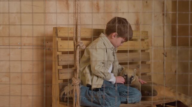 young white boy on wooden pallet inside rustic barn comforting worn teddy bear behind wire mesh, sunlight filtering through slats, straw scattered on floor, patched jacket and denim jeans,