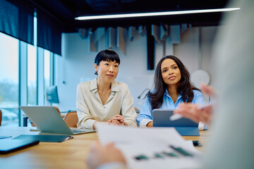 Diverse businesswomen participating in a corporate meeting, listening to a presenter while strategizing and collaborating in the office