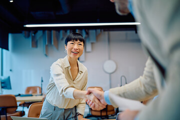 Smiling Asian businesswoman shaking hands with a colleague or client in a modern office, confirming a successful business deal © we.bond.creations