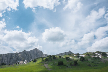 Vivid landscape with green hills with conifer trees and high rock mountain wall under lush clouds in blue sky. Big sharp rocky ridge with sheer crags. Sunlight and shadows in valley in cloudy weather.