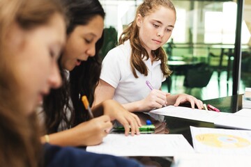 Three young students focused on writing at a table. Diverse group of girls engaged in study. Concentrated students working on assignments together. Education and knowledge.