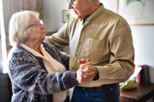 Senior couple dancing in a cozy room. The couple holds hands, sharing a tender moment. The senior pair enjoys a dance, creating a warm, intimate atmosphere. Old couple dancing in retirement home.