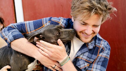 Man with a beard smiling while holding a dog. Dog nuzzles the man. Man and dog share a joyful...