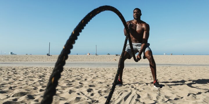 Man exercising on beach with ropes. Fitness on beach, workout with ropes. Beach workout, man using ropes. Exercise on sand, ropes for fitness. African American man doing exercise workout photography.