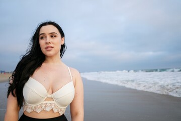 Latina woman with long dark hair stands on a beach, wearing a white top. The ocean waves crash...