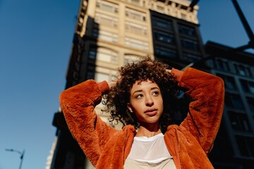 Woman in orange jacket stands in sunlight, urban setting. Curly hair, thoughtful expression. City...