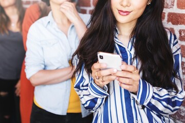 Asian woman using smartphone, standing by a brick wall. Focusing on technology, communication, and modern lifestyle. Asian woman enjoying technology through smartphone.