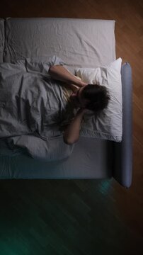 Vertical video man lying in bed awake at night, experiencing insomnia and restless sleep due to stress and anxiety, unable to find comfort or peace in the dark bedroom