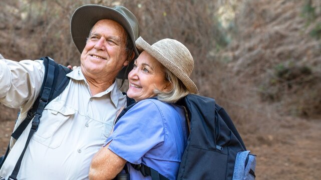 Happy mature couple hiking, wearing hats and backpacks. Smiling seniors enjoying nature. Outdoor adventure for elderly couple, hiking and exploring together. Happy mature couple hiking in nature. - Powered by Adobe