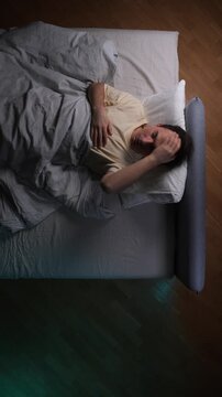 Vertical video man lying in bed at night, covering his face with hands, experiencing sleeplessness and stress, struggling to fall asleep due to anxiety and restless mind, depicting a common problem