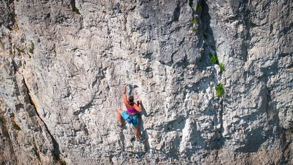 Young adult woman leads a challenging limestone route on a steep outdoor cliff. Fearless climber is belayed with rope and quickdraws. Strength, focus, and skilled movement in a Slovenian climbing area