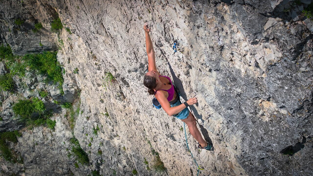 TOP DOWN: Young adult woman stretches for the next hold while lead climbing a steep limestone route. Precision, strength, and authentic motion high above the ground in popular Slovenian climbing area.